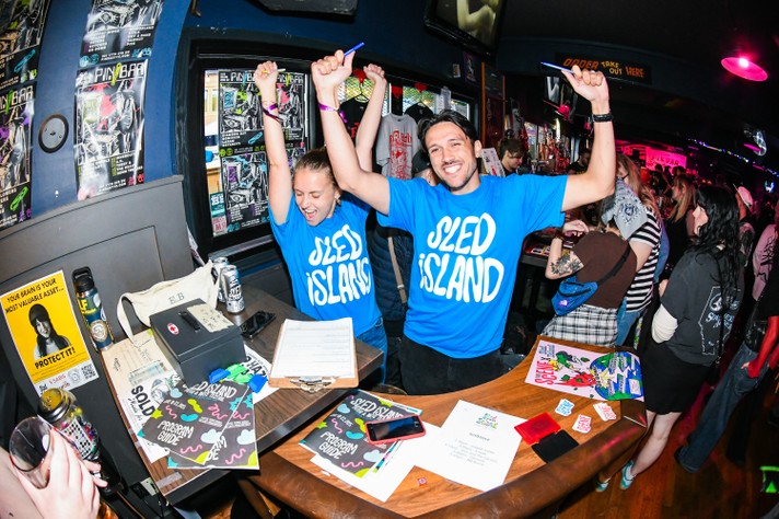A lively scene at a Pin-Bar where two volunteers wearing blue “SLED ISLAND” t-shirts raise their arms in celebration. The counter is cluttered with festival program guides, drink cans, a clipboard, and a bag. Posters decorate the walls, and other patrons chat and mingle in the background, capturing the festive energy of the event.
