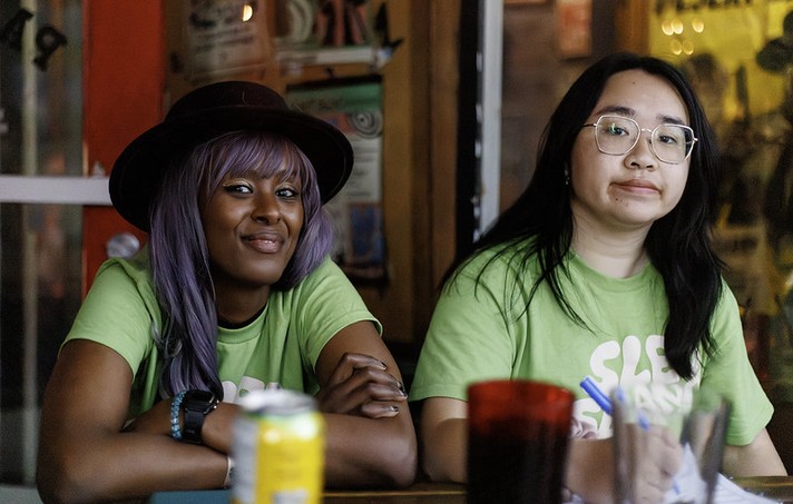 Two people sit side by side at a table indoors, both wearing bright green Sled Island Music & Arts Festival T-shirts. The person on the left has long lavender hair, a dark hat, and a relaxed smile, while the person on the right has long dark hair, glasses, and a neutral expression. On the table in front of them are pens, a red cup, and a yellow drink can. The background shows posters and warm lighting, suggesting a casual, creative festival environment.