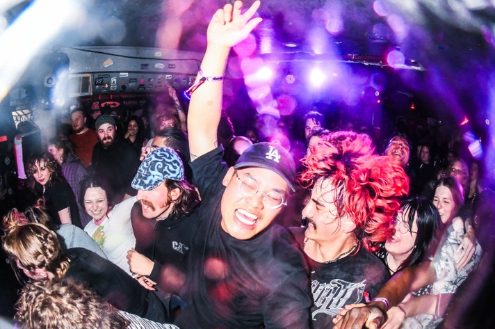 A packed crowd dances and cheers at a live concert in a small, dimly lit venue during Sled Island 2026. In the foreground, a person wearing glasses and a black LA cap raises their arm joyfully, surrounded by others smiling and moving to the music. Another person with bright red hair and a black shirt leans in close, while colorful lights and haze fill the air, creating a vibrant, high-energy atmosphere. The scene captures the excitement and camaraderie of a lively music performance.