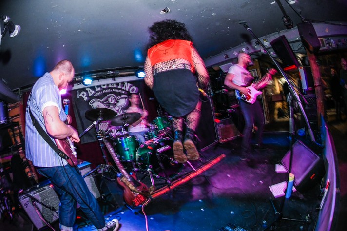 A photo of a high-energy live performance from the band Chinese Medicine at the Palomino during Sled Island 2025, with the lead singer mid-jump, wearing a red top, black skirt, and fishnet stockings. The drummer plays a green Yamaha drum kit, while two guitarists rock out on electric guitars. The stage is filled with musical gear—amplifiers, microphones, and cables. A banner in the background reads “PALOMINO” with a logo, and vibrant stage lighting enhances the dynamic, electrifying atmosphere.