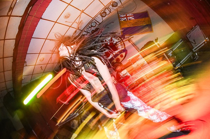 The frontperson of the band afk performs on stage the #1 Legion during Sled Island 2026. Their long dreadlocks is captured mid-motion on stage, their hair flying dramatically as colorful light trails streak across the image. They wear a sleeveless top and patterned pants, leaning forward in an intense pose under bright red, yellow, and green stage lighting. Behind them, musical equipment and a flag hang on the wall, while the blurred motion and vivid colors convey the raw energy and excitement of a live concert.