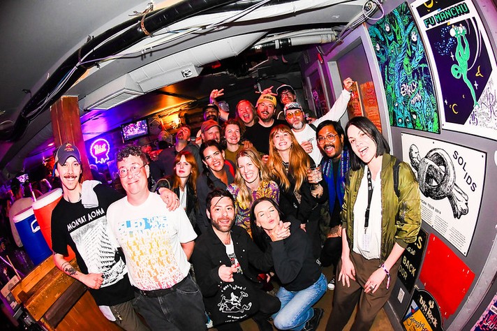 A large group of people gathers inside the Palomino during Sled Island 2026, smiling and posing enthusiastically for the camera. The crowd includes about twenty individuals, some raising their arms or drinks in celebration. Band posters decorate the wall, adding a vibrant, artistic backdrop. The group’s casual clothing and joyful expressions convey a sense of camaraderie and shared excitement, likely after a live show or festival event.