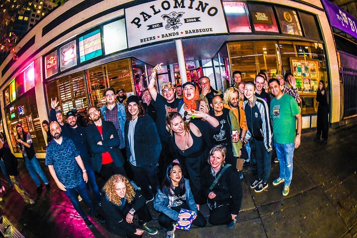 A large, cheerful group of people poses together outside The Palomino Smokehouse in Calgary at night during Sled Island 2026. The restaurant’s sign above them reads “Palomino — Beers, Bands, Barbeque.” The crowd, dressed casually and colorfully, smiles and makes celebratory gestures, with some raising drinks or flashing peace signs. Neon lights from the venue and nearby signs reflect off the wet sidewalk, adding vibrant pink and yellow tones to the lively street scene.