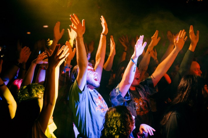 A lively crowd with hands raised in the air under vibrant yellow, blue, and red lighting during a show at Sled Island 2025. The dark background highlights the energetic atmosphere, with attendees dressed in casual clothing like t-shirts and jackets.