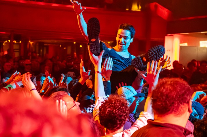 A person is being crowd-surfed at a concert at The Palace Theatre during Sled Island 2025, lifted above a sea of enthusiastic hands. The venue glows with red and orange lighting, amplifying the high-energy atmosphere. The crowd is tightly packed and fully engaged, with the background fading into a dimly lit area filled with more attendees. The person being lifted wears black shoes with distinct tread patterns.