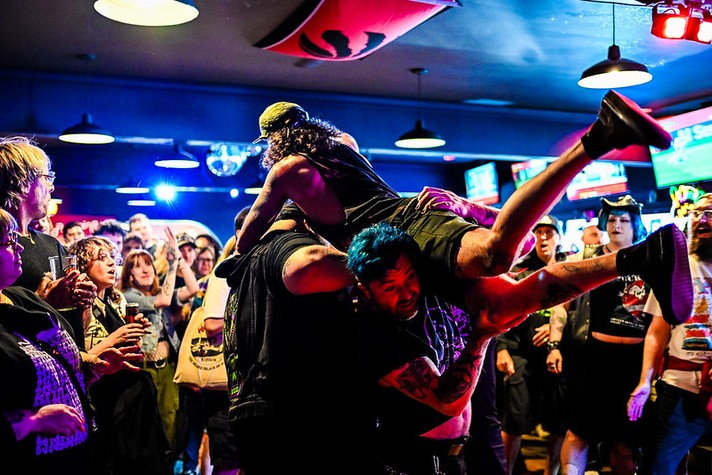 A high-energy moment unfolds at a crowded indoor punk show during Sled Island 2026. In the center, two people lift another person horizontally above their heads, mid-motion, as the crowd cheers and watches closely. The lifted person wears dark clothing and boots, while one of the lifters has bright blue hair and tattooed arms. Colorful stage lights—blue, purple, and red—illuminate the scene, casting vivid reflections across the audience. Around them, people in band shirts and expressive outfits react with excitement, laughter, and awe. Overhead, hanging lamps and a red banner with a black design add to the venue’s lively atmosphere.