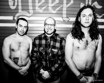 Image description: A black and white press photo for the hardcore trio Show Me the Body, where the three men stand in a line. The bass player and drummer appear shirtless, and the frontman stands in the middle in a plaid jacket, grinning with a cigarette in his mouth.