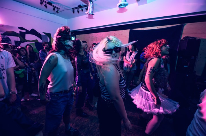 A group of people dance energetically at Loophole during Sled Island 2025, illuminated by purple and blue lights. The crowd features diverse styles, including punk-inspired outfits, tattoos, and distinctive hairstyles. The wooden floor and graffiti-covered wall add to the edgy, underground vibe of the space, capturing a vibrant and expressive moment of music and movement.