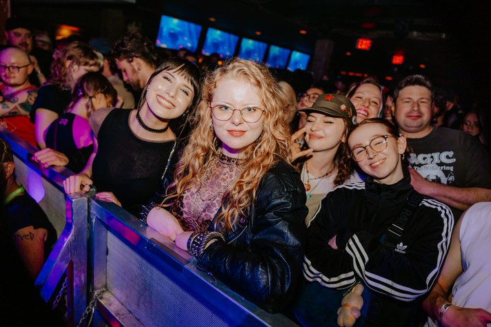 A lively crowd stands at the front of a concert venue during Sled Island 2026, illuminated by colourful stage lights. Several people smile and pose near the barricade, including a woman with short dark hair and a choker, another with long curly blonde hair and glasses wearing a leather jacket, and others behind them making peace signs or grinning. The background shows multiple screens glowing blue and red exit signs above the packed audience, capturing the excitement and camaraderie of a live music event.