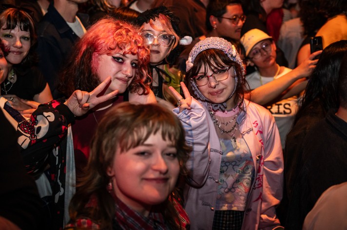 A lively crowd scene at The Palace Theatre during Sled Island 2025 with several people making peace signs. In the foreground, a person wears a light purple jacket with red text on the sleeve, a patterned shirt, and layered necklaces. Another individual to the left sports a black shirt with a colorful patterned sleeve. The background is densely packed with people, creating a vibrant and energetic atmosphere.