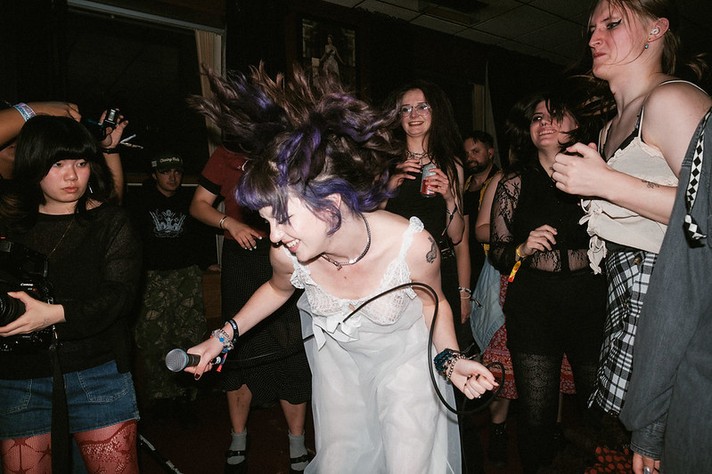 During Sled Island 2026, a performer with purple hair from the band Poolgirl bends forward mid‑motion, holding a microphone while headbanging energetically in front of a lively crowd. They wear a white lace dress and several bracelets, their hair flying as people around her cheer and dance. Audience members in dark clothing, fishnets, and plaid skirts smile and move to the music, with one person holding a camera to capture the moment. The dimly lit venue and close crowd create an intimate, high‑energy atmosphere typical of a small punk or alternative show.