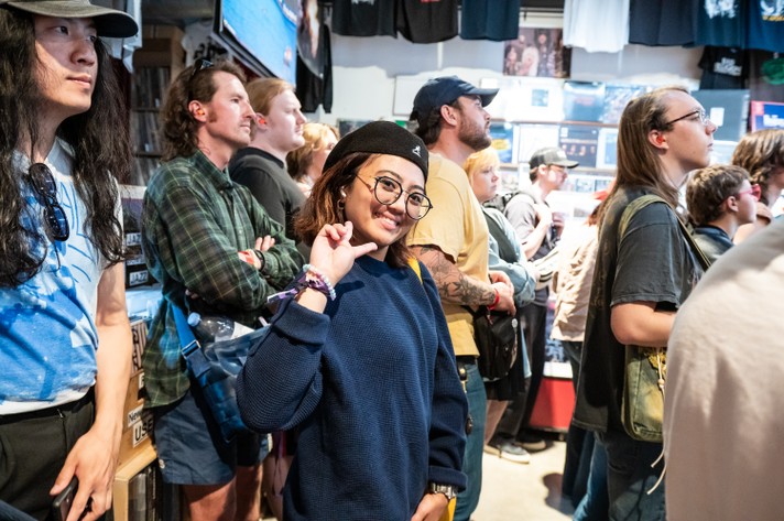 A group of people gather in a crowded indoor space at Sloth Records for a show during Sled Island 2025. The walls are decorated with merchandise like t-shirts and posters. In the foreground, a person wearing a blue sweater and black cap holds up their fingers to make a peace sign. Others around them are casually dressed, some with hats and bags, contributing to the lively and social atmosphere.