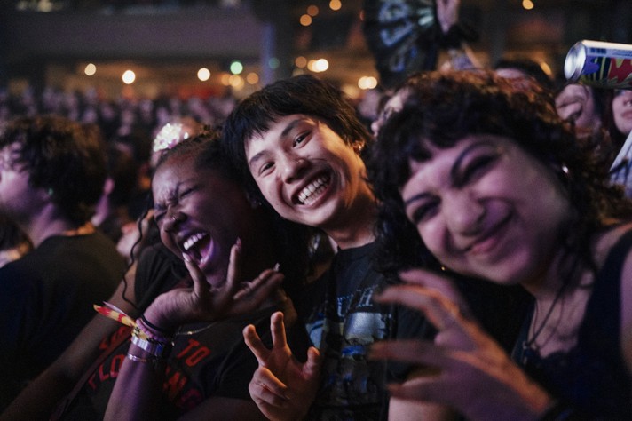 During Sled Island 2026, three friends stand at the front of a crowded concert, laughing and posing for the camera. The person on the left throws their head back with a joyful expression, wearing colorful wristbands. The person in the middle grins widely, flashing a peace sign, while the person on the right scrunches their face playfully and raises their hands like claws. Behind them, a packed audience glows under warm stage lights, capturing the excitement and energy of a live music event.