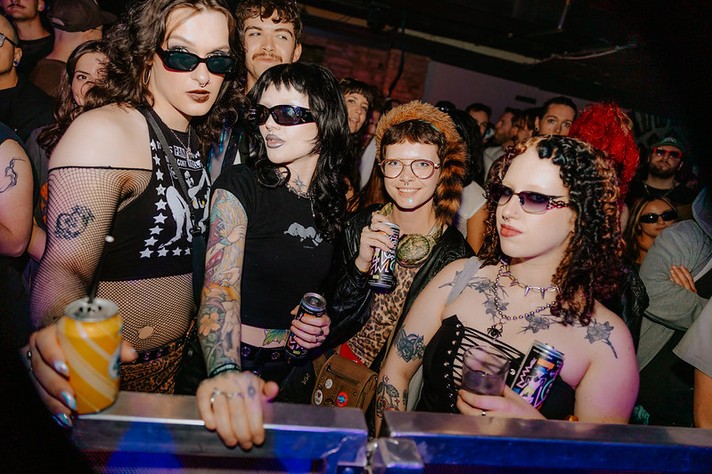During Sled Island 2026, a group of friends stands at the front of a crowded concert venue, illuminated by colorful stage lights. They wear bold, expressive outfits featuring mesh tops, tattoos, sunglasses, and studded accessories, each holding a drink can or cup. The person on the left wears dark lipstick and a fishnet shirt with a star pattern, while others sport black clothing, animal‑print details, and layered jewelry. Behind them, the audience fills the space, watching the stage with excitement, capturing the vibrant energy and individuality of the live music scene.
