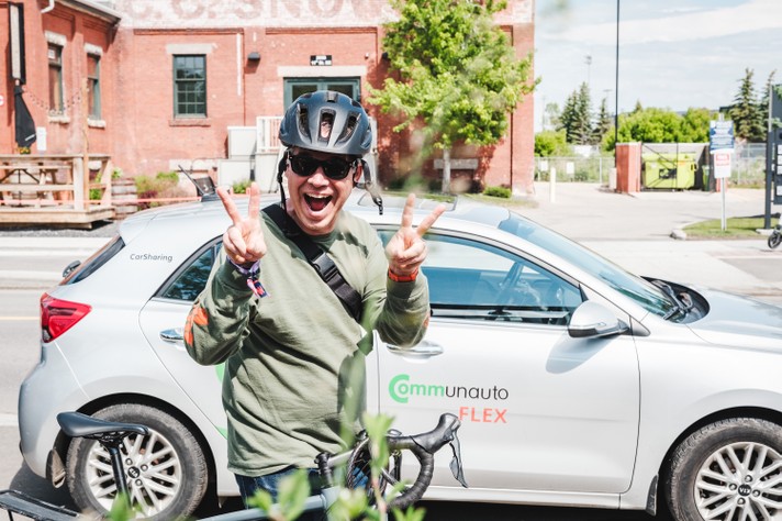 During Sled Island 2026, a smiling person wearing a black bike helmet and sunglasses flashes double peace signs while standing beside a silver car with “Communauto FLEX” and “CarSharing” logos. They wear a green long-sleeve shirt and have a black shoulder strap across their chest. A bicycle is visible in the foreground, and behind them stands a red brick building with a faded painted sign. The bright, sunny scene conveys a cheerful, eco-friendly vibe centered on urban mobility and shared transportation