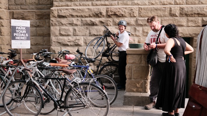 A group of bicycles parked in a designated area beside the Central United Church during Sled Island 2025. A sign reads “PARK YOUR PEDALS HERE!” with smaller text below. One person lifts a bicycle, while two others chat nearby. The scene conveys a communal, bike-friendly space for secure parking and social interaction.