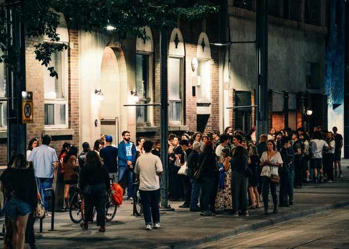 A large crowd gathers outside Central United Church during Sled Island 2025, featuring arched windows and warm lighting. People stand and sit on bicycles, chatting and socializing. A “No Parking” sign is visible on a nearby pole, and the scene is lit by streetlights and building lights, creating a lively, communal atmosphere.