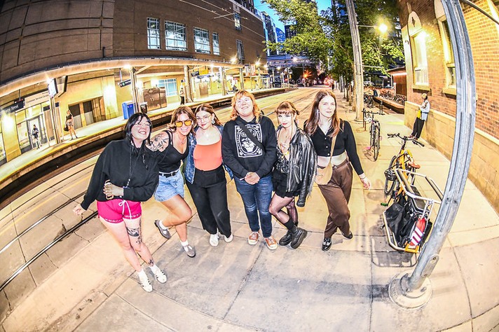 During Sled Island 2026, seven people stand together on a city sidewalk near a c-train station at dusk, smiling and posing for the camera. The photo, taken with a fisheye lens, curves the edges of the scene for a dynamic effect. The group wears casual outfits—shorts, jeans, jackets, and sneakers—and some have brightly colored glasses or tattoos. Behind them, the station platform glows under streetlights, with a few people waiting and bicycles parked along the brick building to the right. The atmosphere feels friendly and urban, capturing a lively evening moment.
