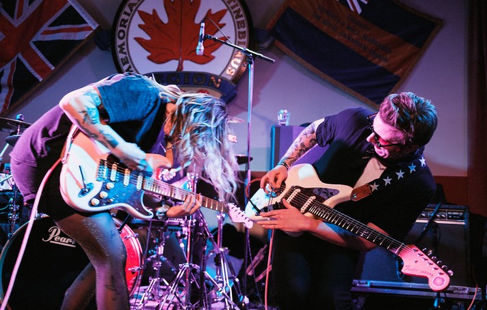 During Sled Island, two guitarists from the group Sub / / Cell perform energetically on stage, leaning toward each other as they play electric guitars under colorful purple and blue lights. Both musicians are mid-motion, their hair and posture reflecting the intensity of the moment. Behind them, a drum set and flags—including a Union Jack and a Canadian Legion emblem featuring a red maple leaf—decorate the venue. The scene captures the raw power and camaraderie of a live rock performance.