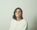 Indoor portrait of solo artist Rae Spoon smiling toward the camera, in front of a neutral background.