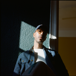 Portrait of rapper Oddisee seated and looking down toward the camera, with the window simultaneously illuminating and casting a shadow on him.