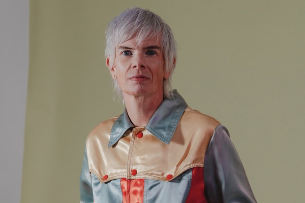Indoor portrait of solo artist Rae Spoon, looking toward the camera in front of a neutral background.