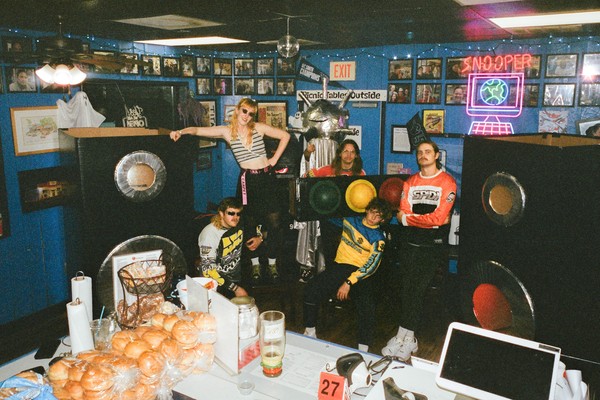 Six members of Snõõper are posed between two large speakers. One is costumed, wearing a spiky silver headdress on top of fabric disguising their body.