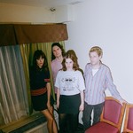 Indoor portrait of four-piece TOPS standing in the corner of a room, between two chairs and a window.
