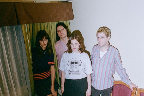 Indoor portrait of four-piece TOPS standing in the corner of a room, between two chairs and a window.