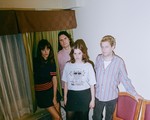 Indoor portrait of four-piece TOPS standing in the corner of a room, between two chairs and a window.
