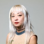 Artist Evicshen is shown posing for a colour photo in front of an off-white background. They are shown from the shoulders up, wearing a tan shirt and having bleach blonde shoulder length hair