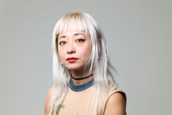 Artist Evicshen is shown posing for a colour photo in front of an off-white background. They are shown from the shoulders up, wearing a tan shirt and having bleach blonde shoulder length hair