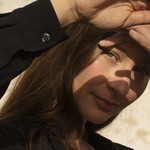 A close-up colour photo shows Kara-Lis Coverdale with their hands against their face blocking sunlight from their eyes. They are wearing a dark long sleeved shirt. A beige wall can be seen in the background.
