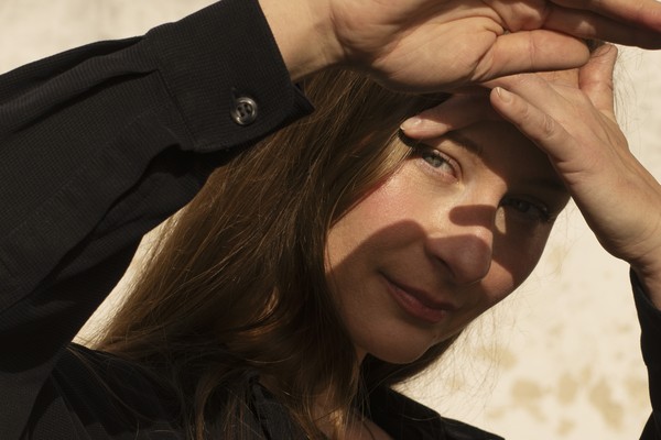 A close-up colour photo shows Kara-Lis Coverdale with their hands against their face blocking sunlight from their eyes. They are wearing a dark long sleeved shirt. A beige wall can be seen in the background.