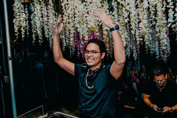 DJ BLKFT is seen performing with headphone around their neck, raising their hands in the air enthusiastically. White flowers hang behind them. 