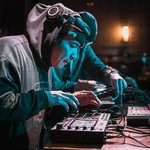 Artist Creeasian is seen hunched over some DJ gear while performing live. They are wearing a black sweater with the hood up, and their headphone on over top.