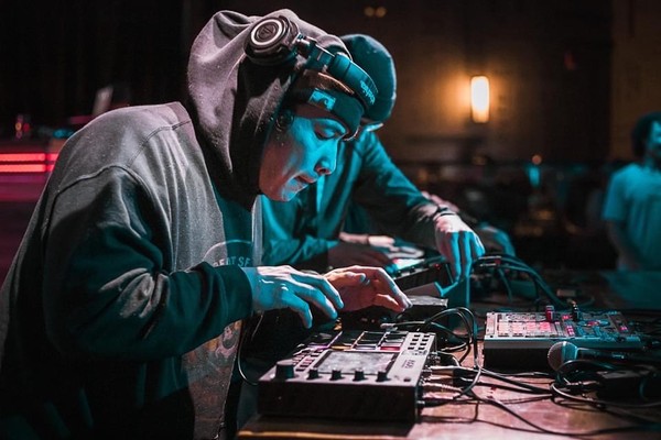 Artist Creeasian is seen hunched over some DJ gear while performing live. They are wearing a black sweater with the hood up, and their headphone on over top.