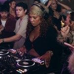 A colour photo of DJ Blexxy as she is intensely focused while mixing music on a DJ controller at a crowded party. She is wearing a leopard print headscarf and headphones. She is closely surrounded by a diverse group of people dancing.