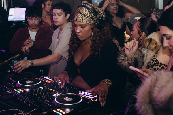 A colour photo of DJ Blexxy as she is intensely focused while mixing music on a DJ controller at a crowded party. She is wearing a leopard print headscarf and headphones. She is closely surrounded by a diverse group of people dancing.