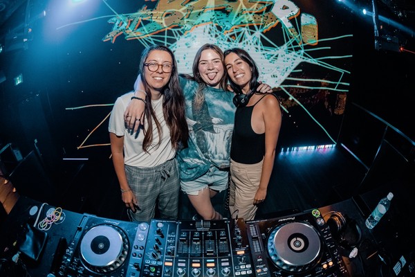 The three members of the DJ group LVL Collective pose for a colour photo standing on stage behind a turntable and DJ setup. 