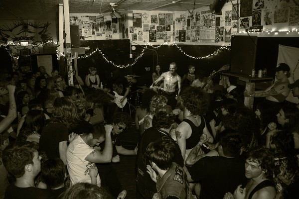 A black and white photo shows the band SMUTHER playing a packed intimate show, standing in the corner of a small room. 