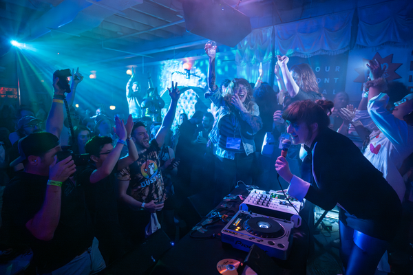 A colour photo shows Annie-Claude Deschanes playing an intimate show in front of a very enthusiastic crowd. They are seen singing into a microphone, behind a turntable. Everyone is bathed in blue light. 