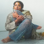 A colour photo shows artist Thanya Iyer sitting cross-legged in a white room tucking their right knee up to their face. They are wearing knitted net-like pants and have various flower petals stuck to their face to resemble colourful face paint. 
