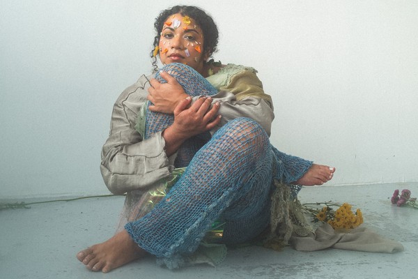 A colour photo shows artist Thanya Iyer sitting cross-legged in a white room tucking their right knee up to their face. They are wearing knitted net-like pants and have various flower petals stuck to their face to resemble colourful face paint. 