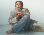 A colour photo shows artist Thanya Iyer sitting cross-legged in a white room tucking their right knee up to their face. They are wearing knitted net-like pants and have various flower petals stuck to their face to resemble colourful face paint. 