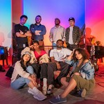 A colour photo of the band (un)decided), featuring all eight members posing in a warmly lit studio space with vibrant red, blue, and purple lighting. Four members sit on the floor or couch in front while the other four stand behind them. The space is filled with musical equipment, including guitars, drums, and keyboards.