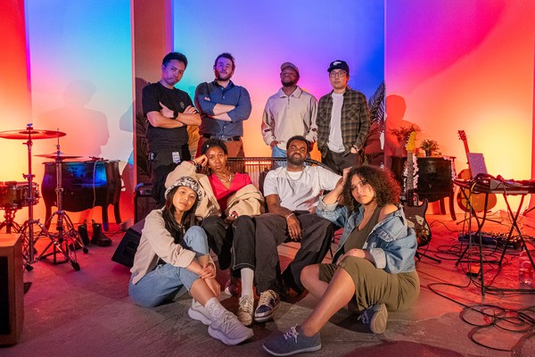 A colour photo of the band (un)decided), featuring all eight members posing in a warmly lit studio space with vibrant red, blue, and purple lighting. Four members sit on the floor or couch in front while the other four stand behind them. The space is filled with musical equipment, including guitars, drums, and keyboards.