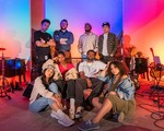 A colour photo of the band (un)decided), featuring all eight members posing in a warmly lit studio space with vibrant red, blue, and purple lighting. Four members sit on the floor or couch in front while the other four stand behind them. The space is filled with musical equipment, including guitars, drums, and keyboards.