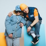 Vibrant studio portrait of duo 538st. One is seated on a blue block and the other is standing in front of an orange block.
