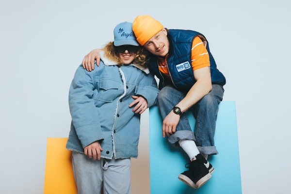 Vibrant studio portrait of duo 538st. One is seated on a blue block and the other is standing in front of an orange block.
